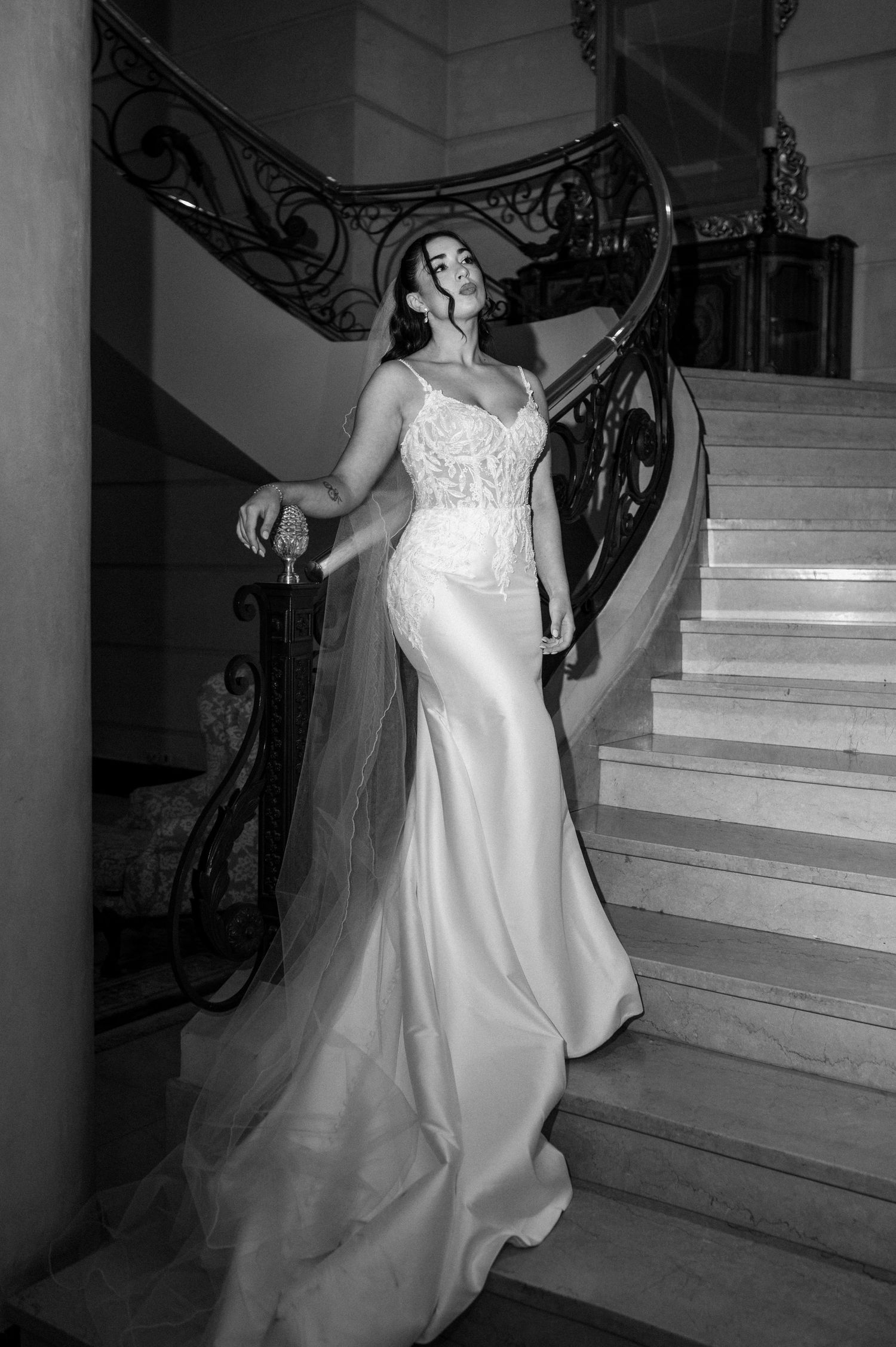 Novia con un elegante vestido de novia de encaje estilo sirena posa en una gran escalera con una barandilla de hierro ornamentada, fotografí