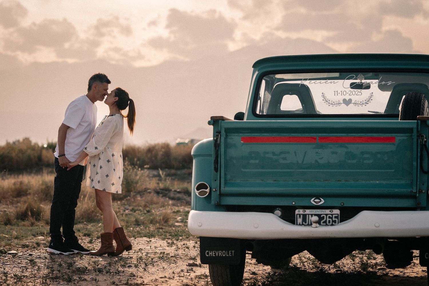 A couple shares a romantic moment at sunset near a vintage teal pickup truck in a desert landscape.
