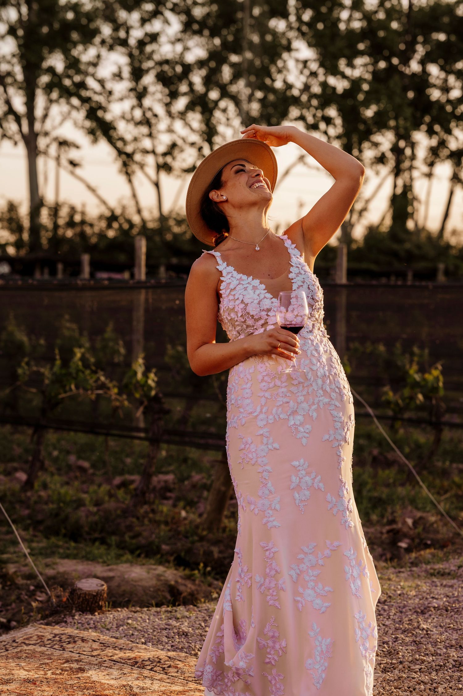 Woman in a flowing lace wedding dress and sun hat poses in a vineyard at golden hour.