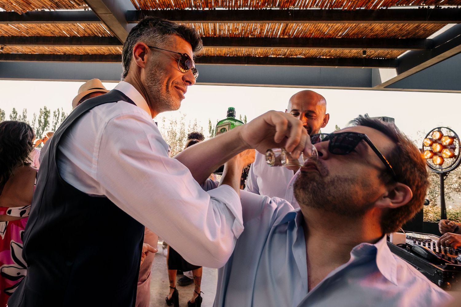 Wedding guests share celebratory moments during an outdoor reception under a rustic wooden canopy.