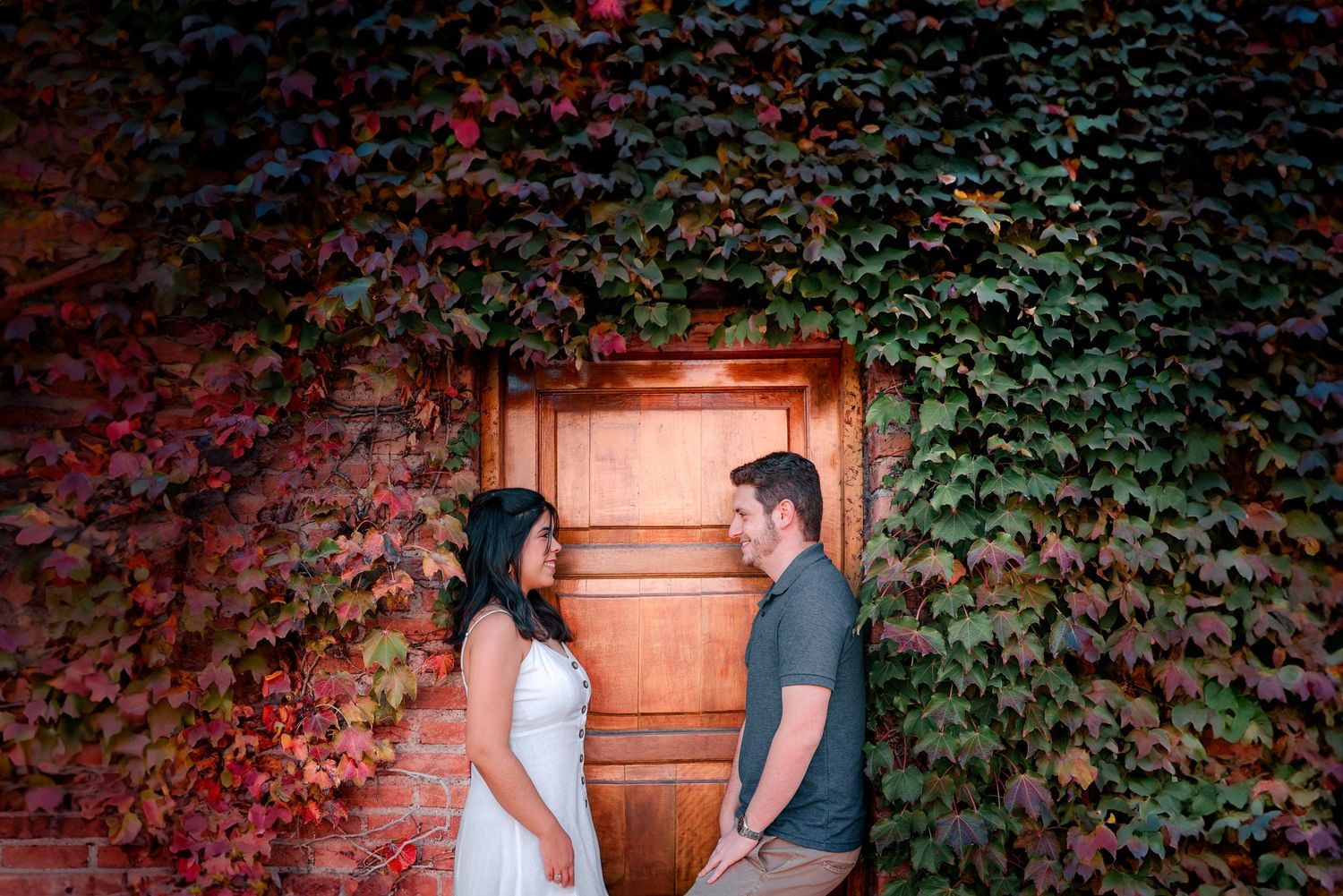 A couple stands before a rustic wooden door surrounded by lush green ivy and fall foliage for a romantic photoshoot.