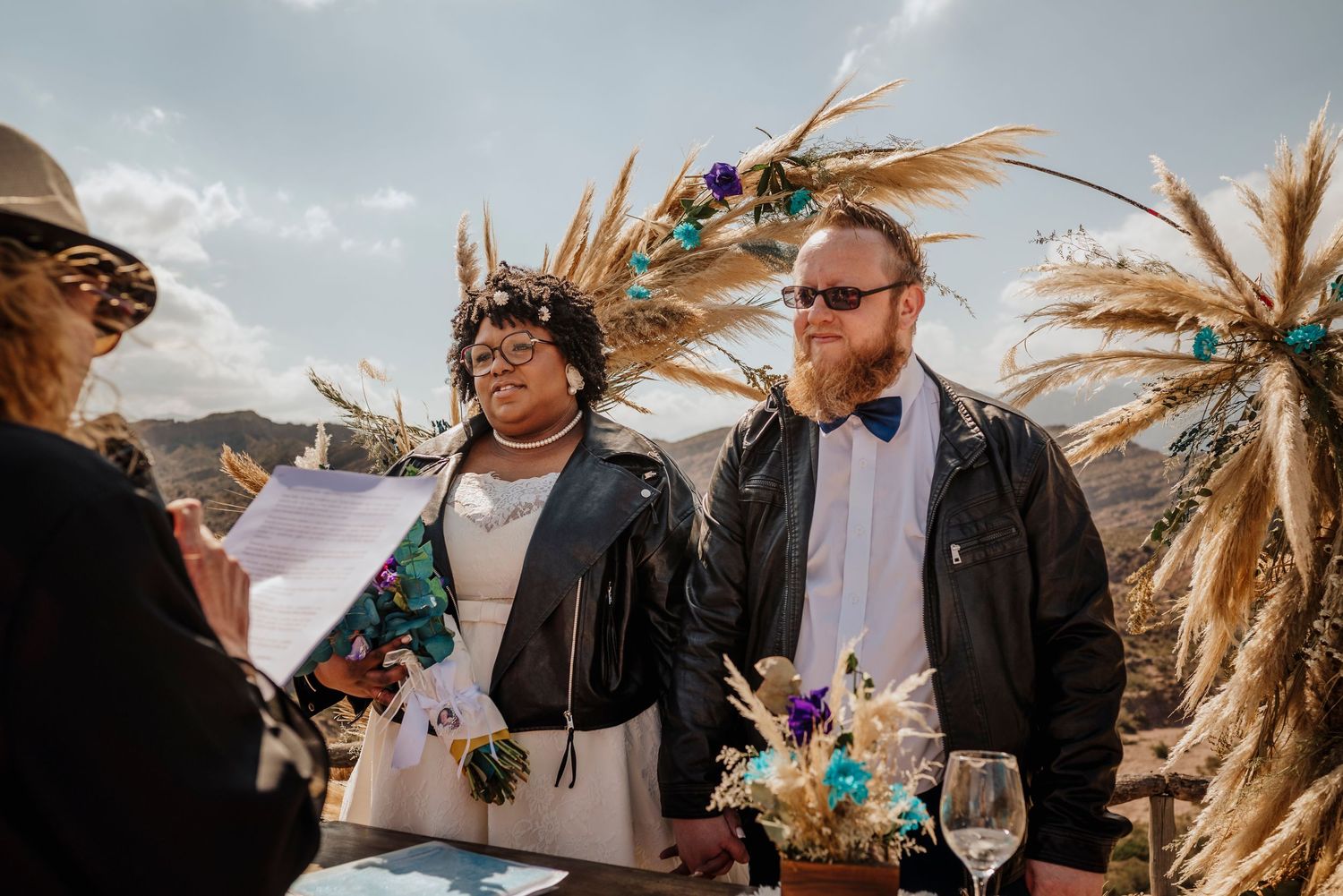 Wedding ceremony in a rustic outdoor setting with tall pampas grass decorations against a cloudy sky.