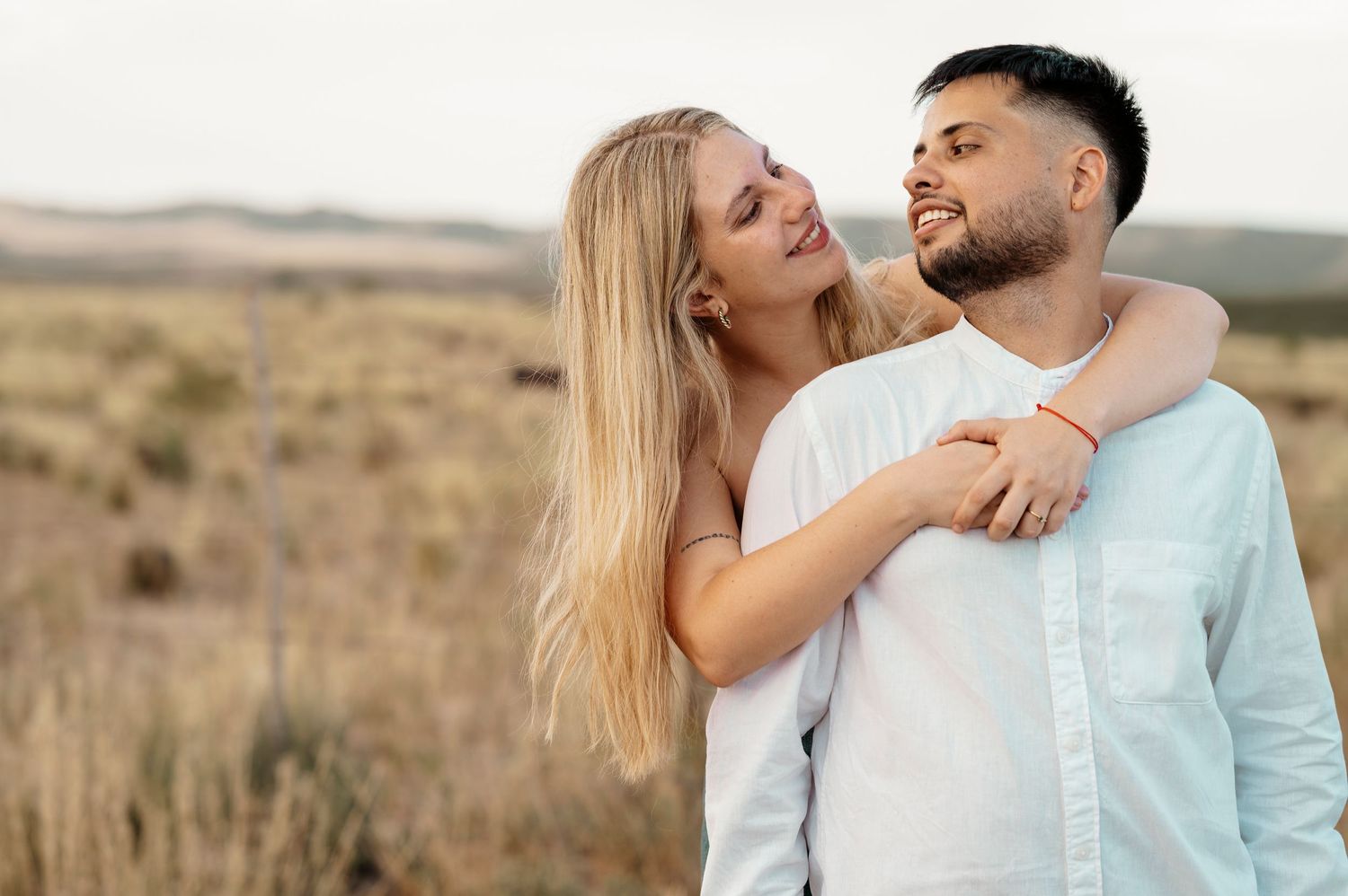 A couple in light colored clothing embraces and laughs together in a scenic desert landscape setting.