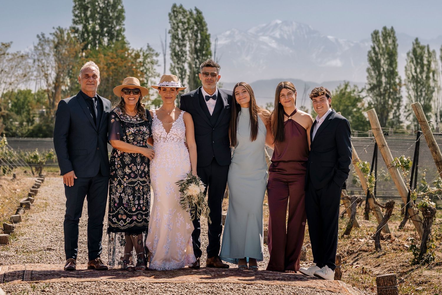 A group of wedding attendees pose together outdoors against a scenic mountain backdrop on a sunny day.