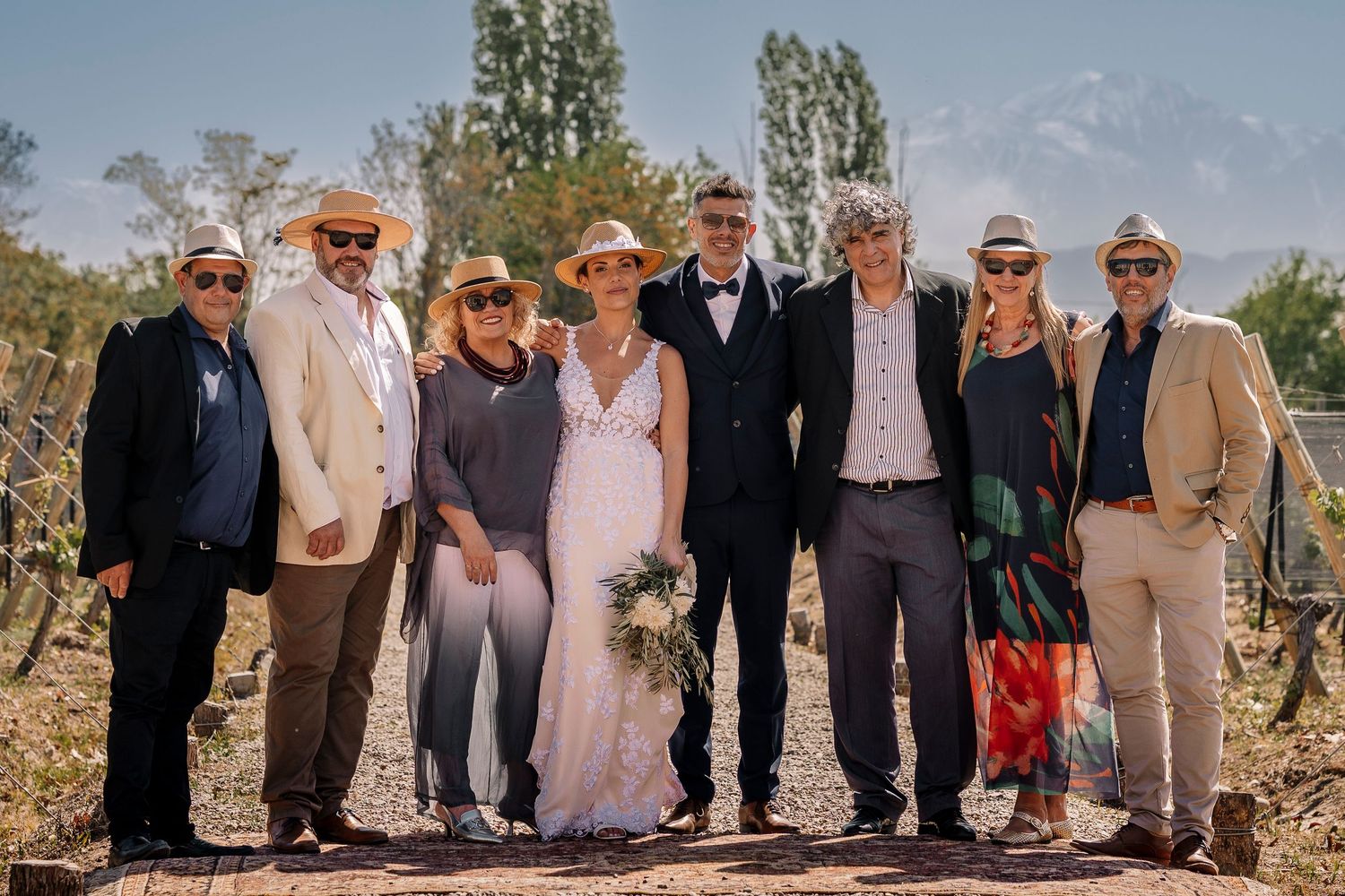A group of wedding guests pose together outdoors in a sunny vineyard setting with trees and mountains in the background.