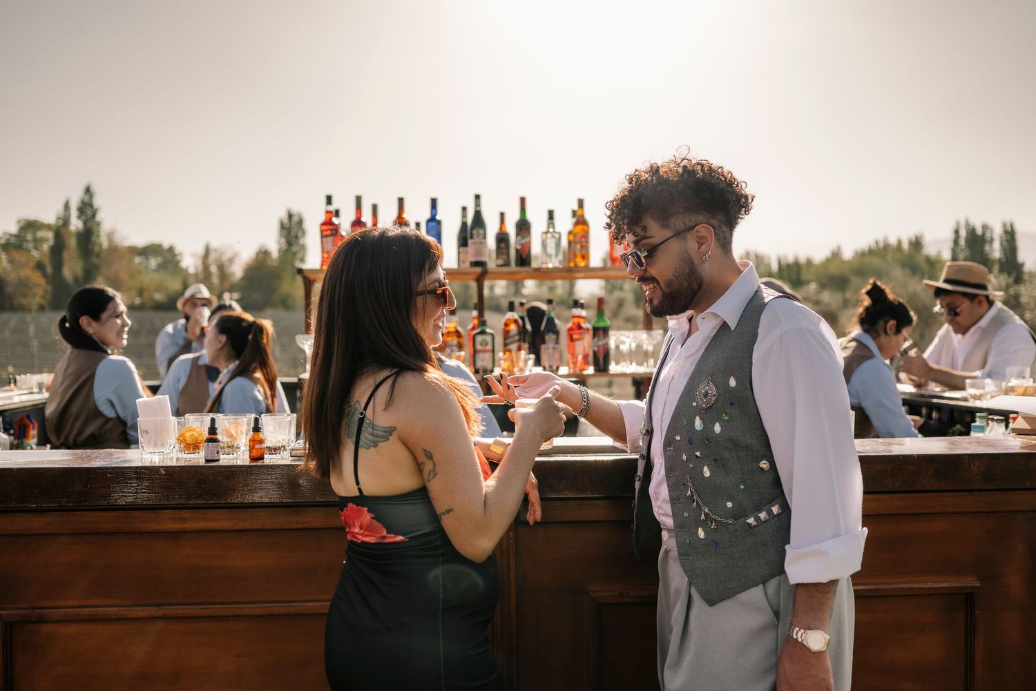 People enjoying drinks at an outdoor bar with liquor bottles displayed on shelves during a sunny afternoon gathering.