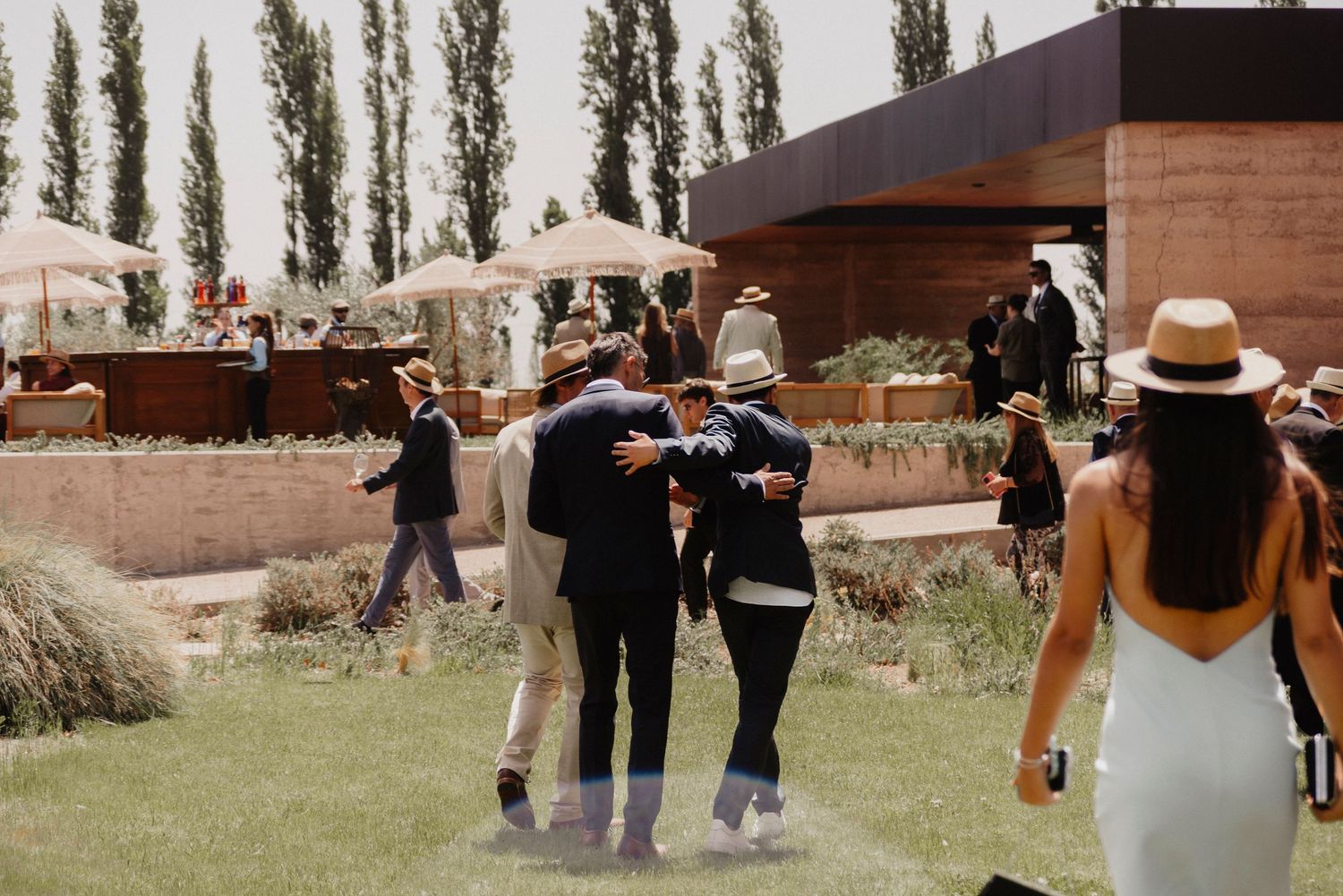 A wedding party walks through a garden venue with tall poplar trees in the background.