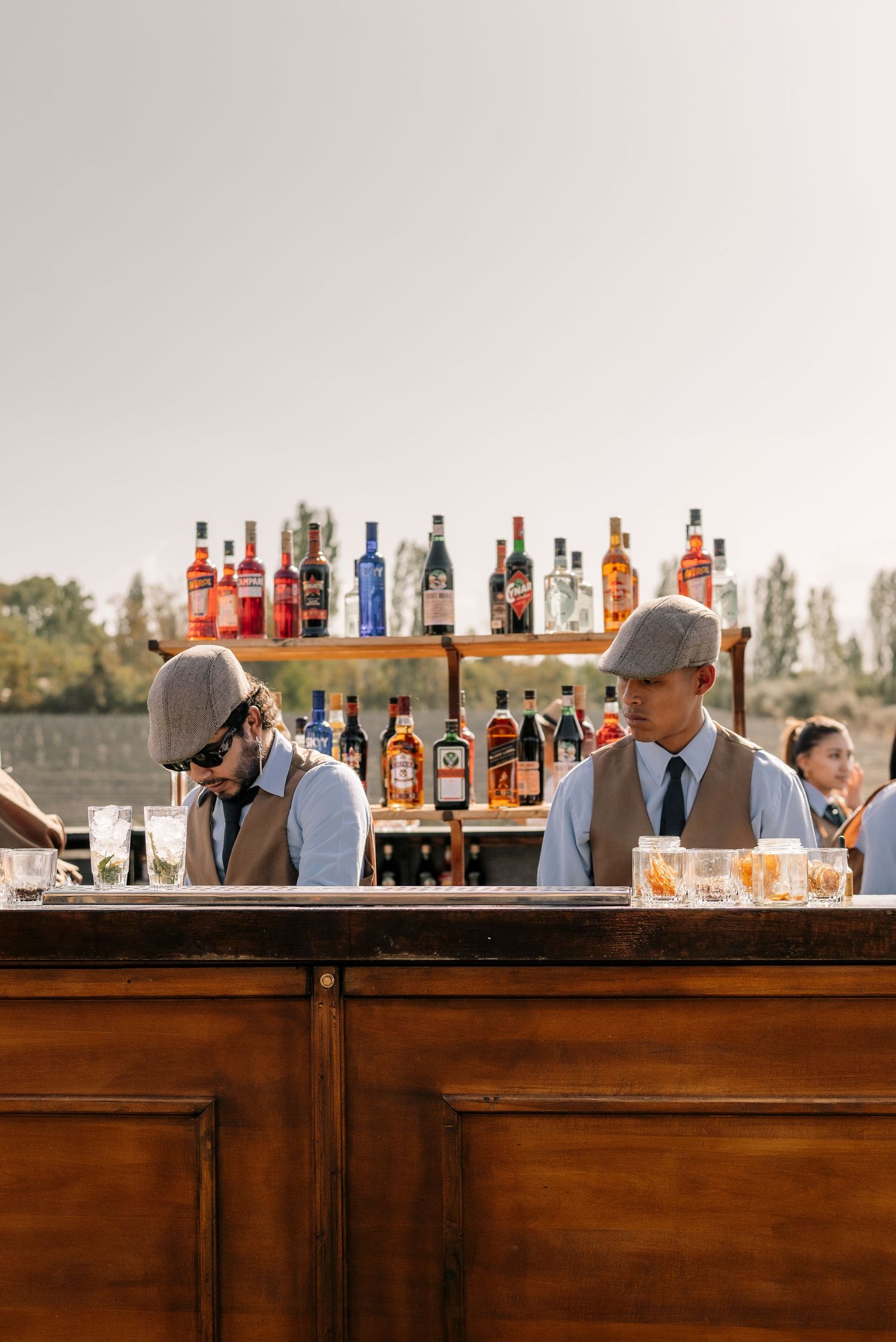 Wooden bar setup with rows of liquor bottles and bartenders serving drinks at an outdoor event.