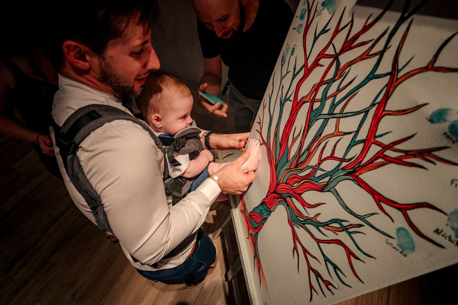 Baby in carrier helps add fingerprint leaves to a decorative tree painting during a creative family activity.