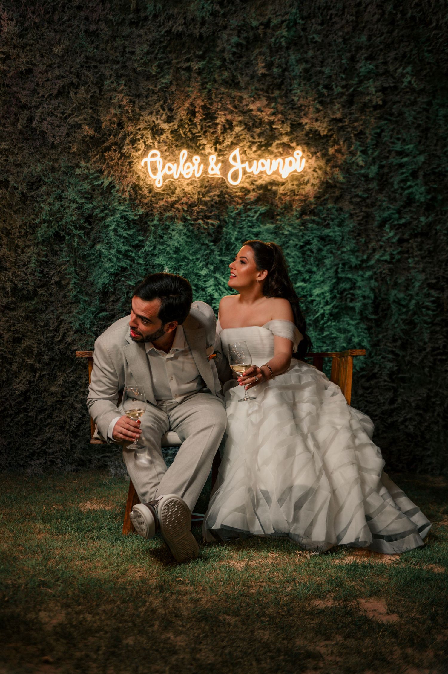 Romantic wedding couple sits on grass under glowing neon sign at dusk during outdoor reception.