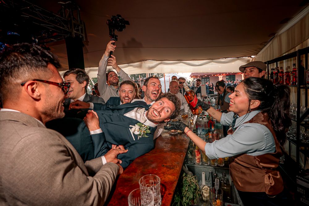 A group of people cheering and raising drinks together in celebration at a crowded bar.