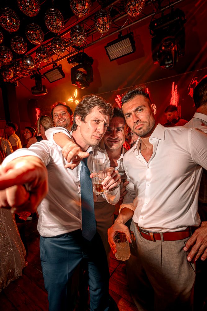 Wedding party guests dancing and celebrating in a dimly lit reception venue with red lighting.