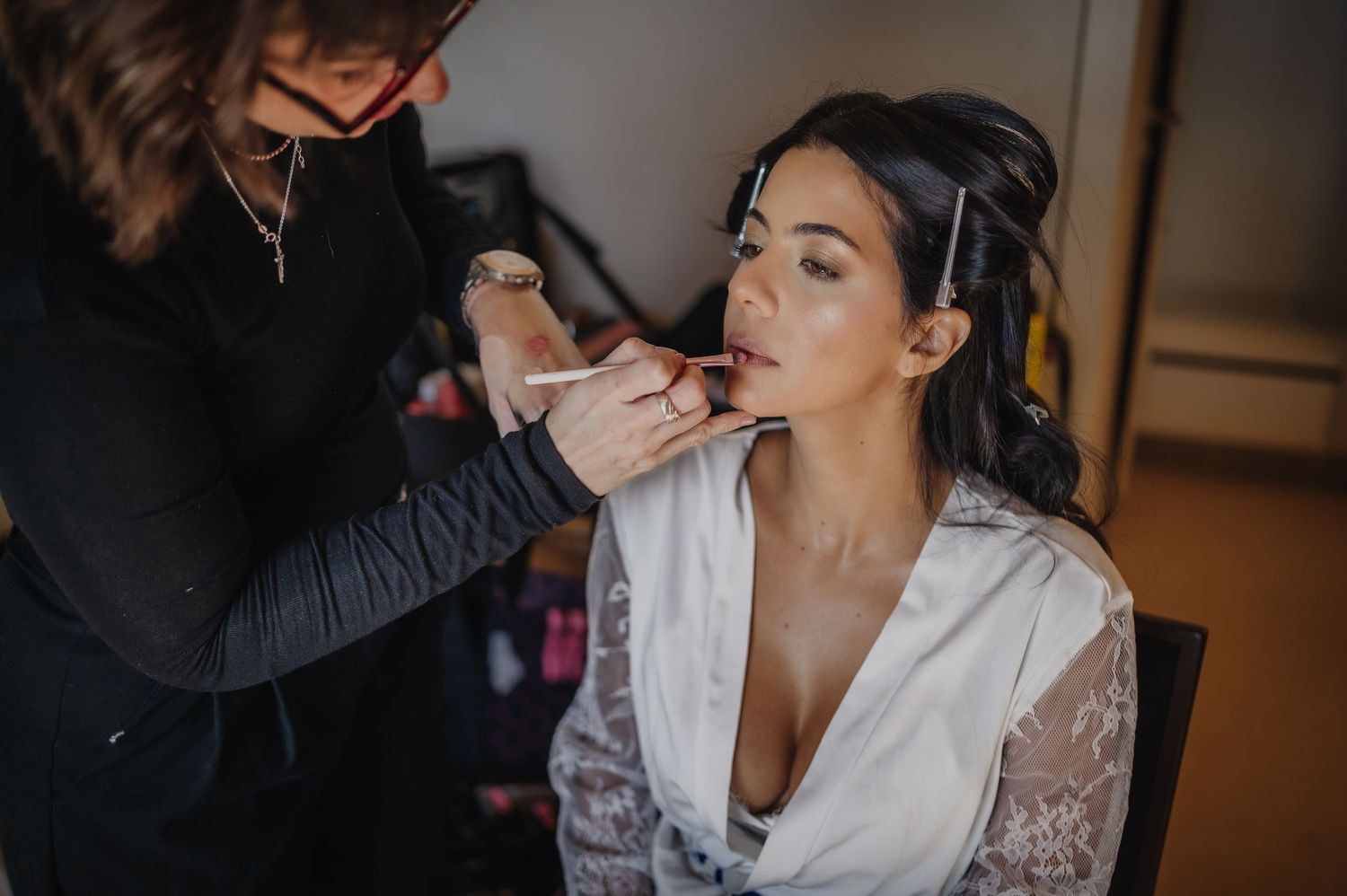 Makeup artist applying lipstick to a bride in a white lace robe during wedding day preparations.