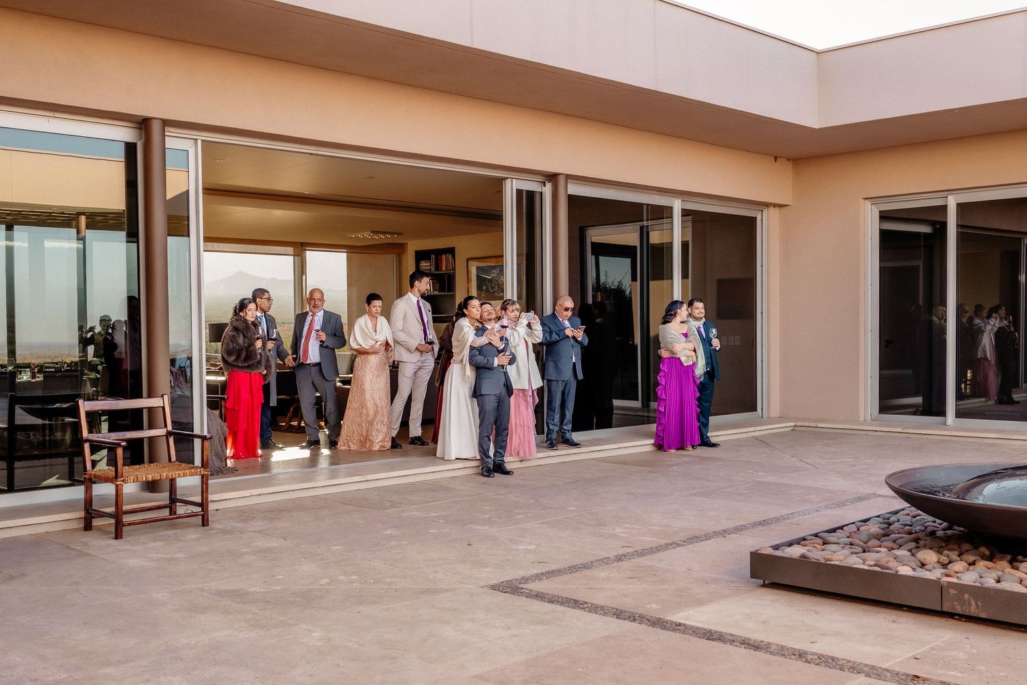 Wedding guests gathered on a sunlit patio outside modern venue with large glass doors during an outdoor celebration.