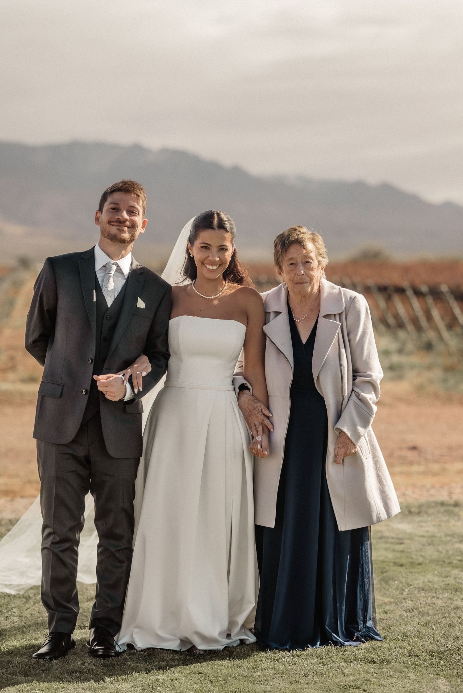 A bride and groom pose with an older woman in a vineyard setting with mountains in the background.