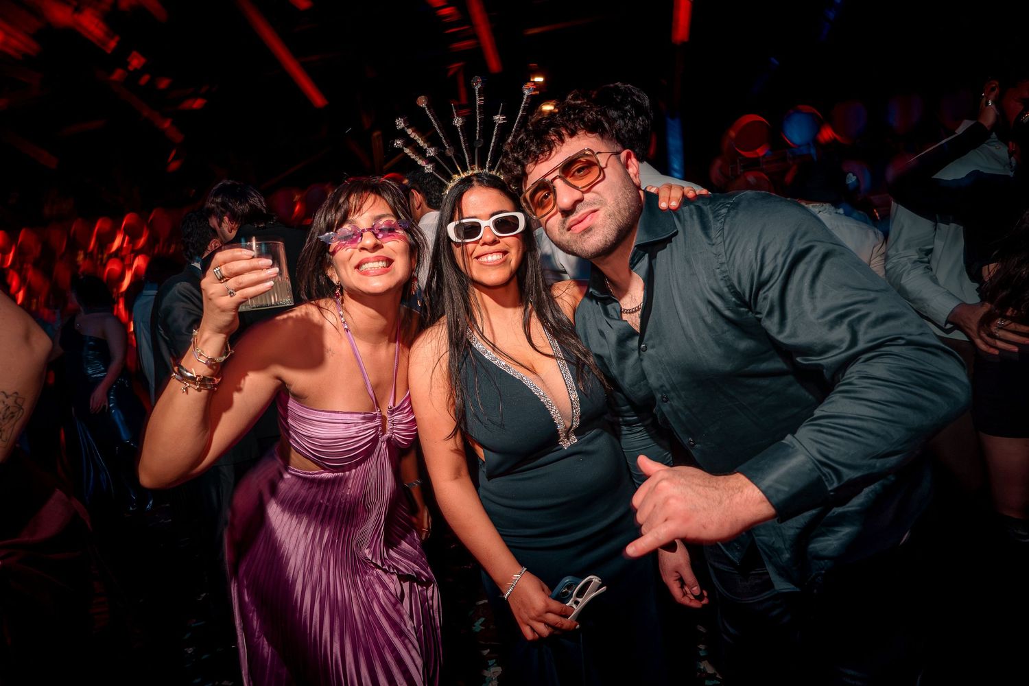 Three friends posing together at a nightclub party, wearing festive accessories and stylish outfits under colorful lights.