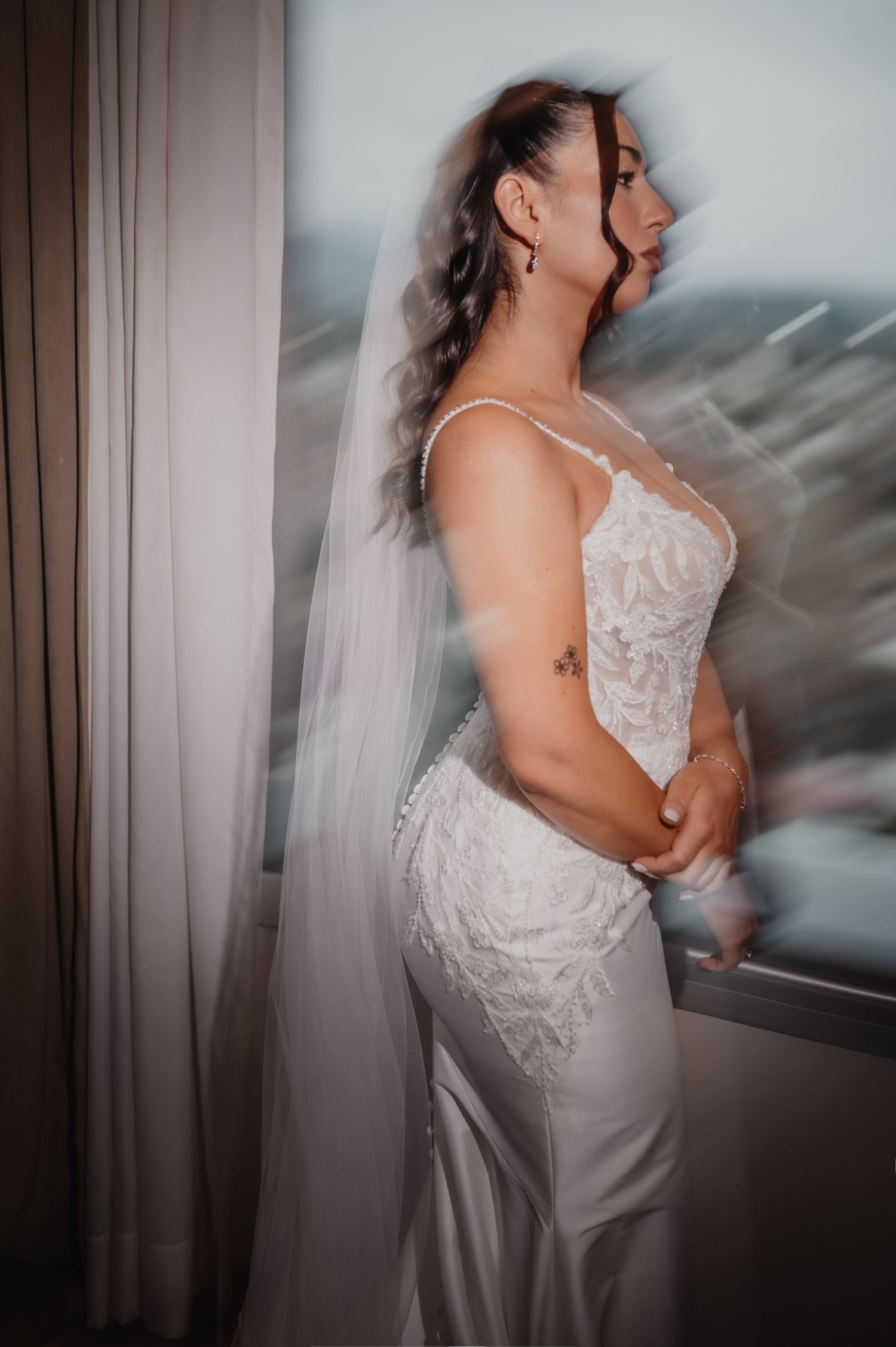 La novia, con vestido de novia de encaje blanco y velo, posa junto a la ventana, contemplando el exterior en un elegante retrato nupcial.