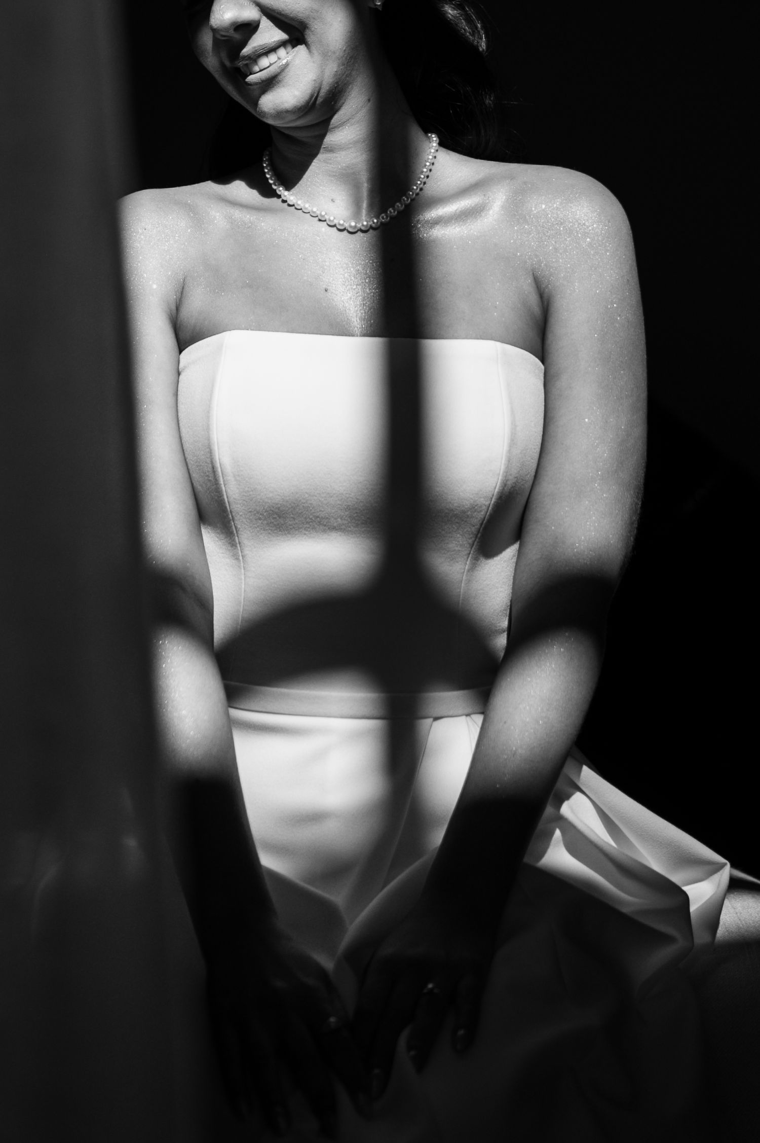 Black and white photo of a smiling bride in a strapless gown with pearl necklace, dramatic light shadows across her dress.