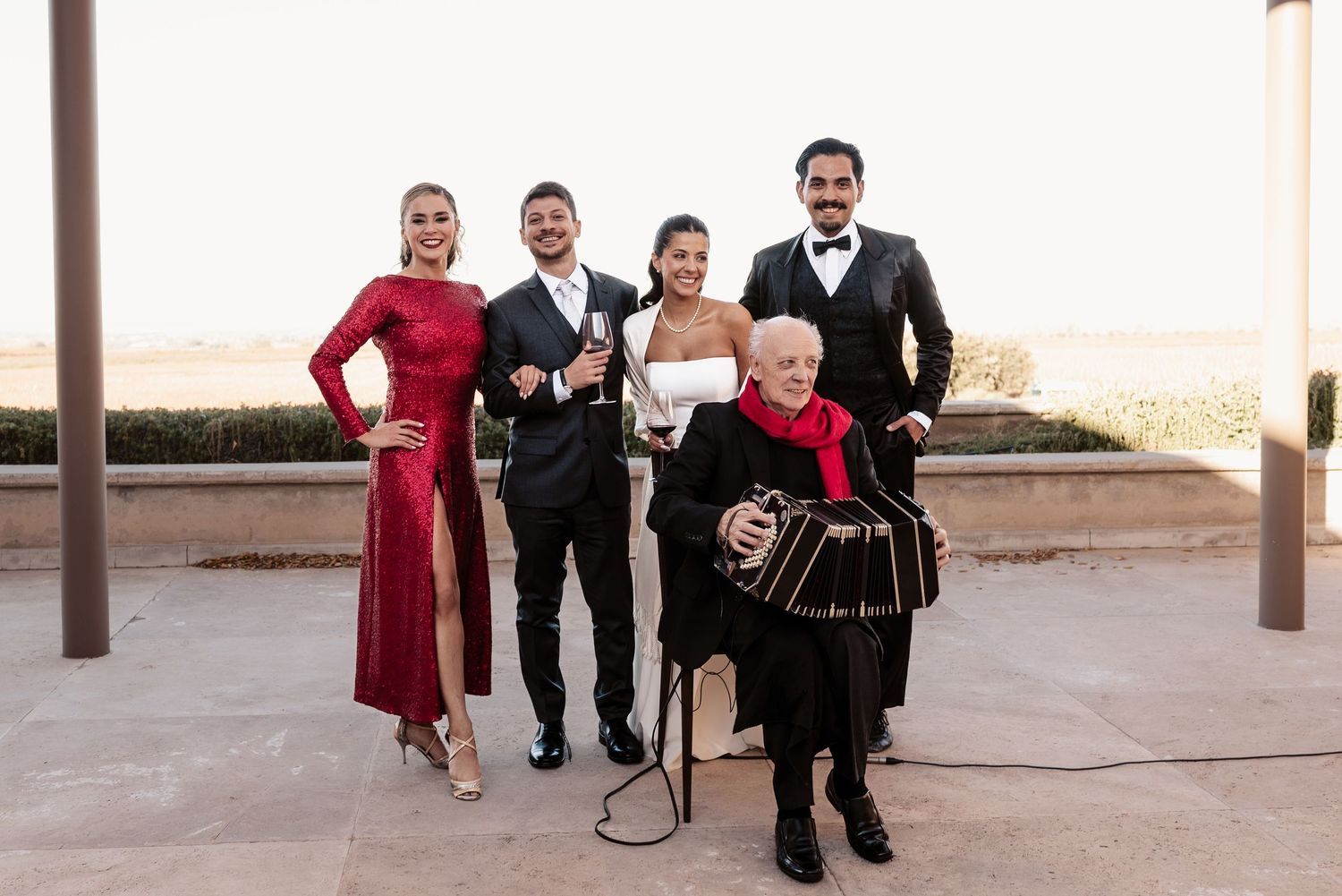 Five elegantly dressed performers pose outdoors, including a woman in red, a musician with an accordion, and three others in formal attire.