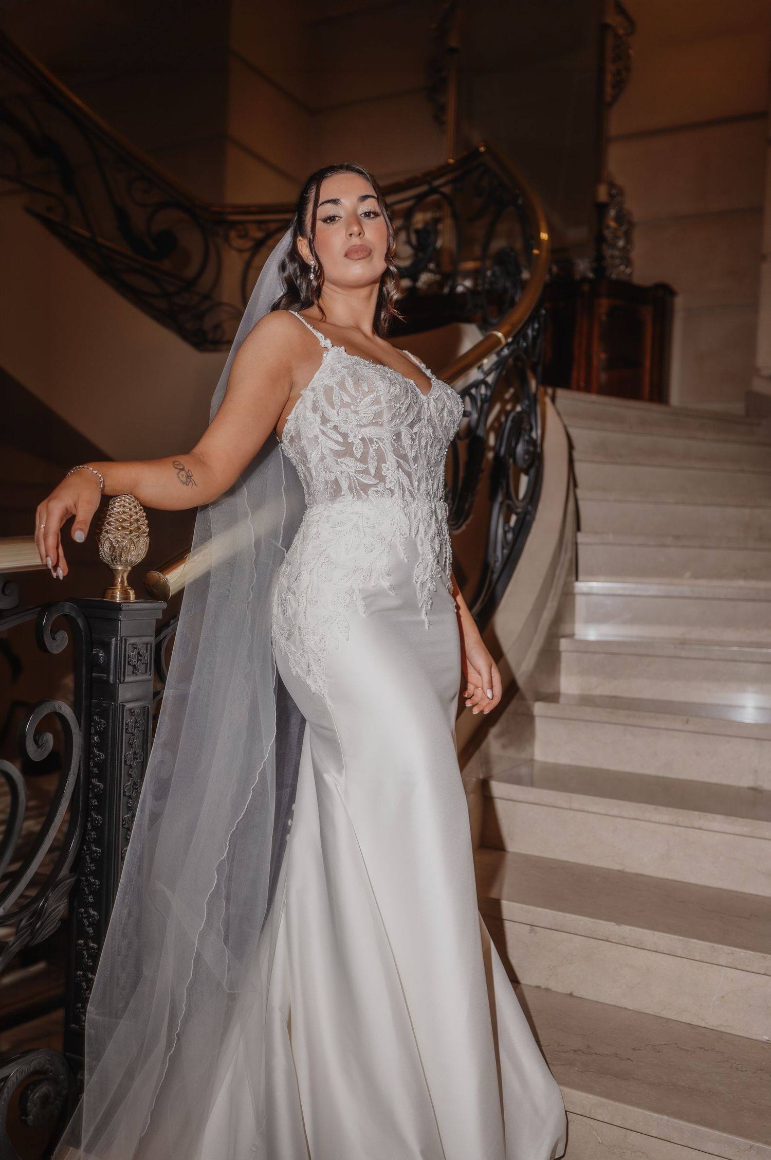 Bride in lace mermaid wedding gown with veil poses on elegant marble staircase with ornate iron railings.