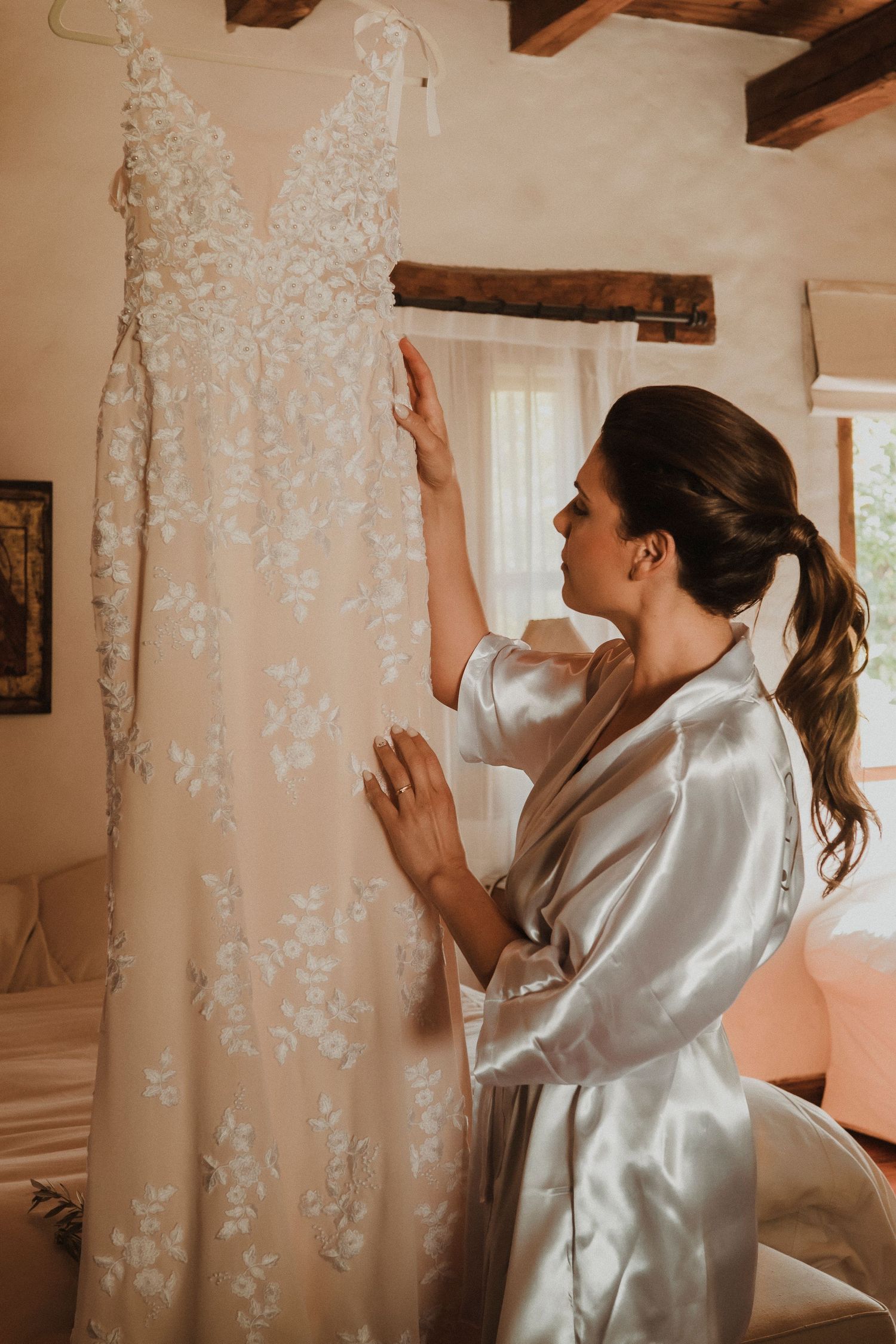 A person in a silky white robe examines delicate lace details on a hanging garment in a rustic room with wooden beams.
