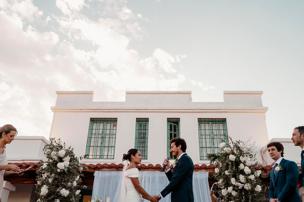 A wedding couple and guests celebrate during an outdoor reception at sunset with a white building in the background.