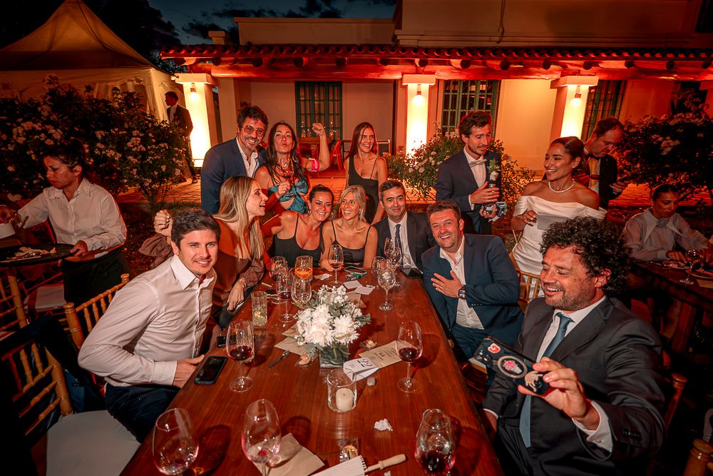 Wedding guests gather around a warmly lit table at a reception dinner with glowing orange lanterns in the background.