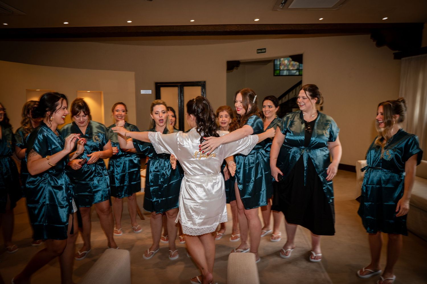 A group in matching teal robes surrounds someone in white during a celebratory dance moment at an indoor venue.