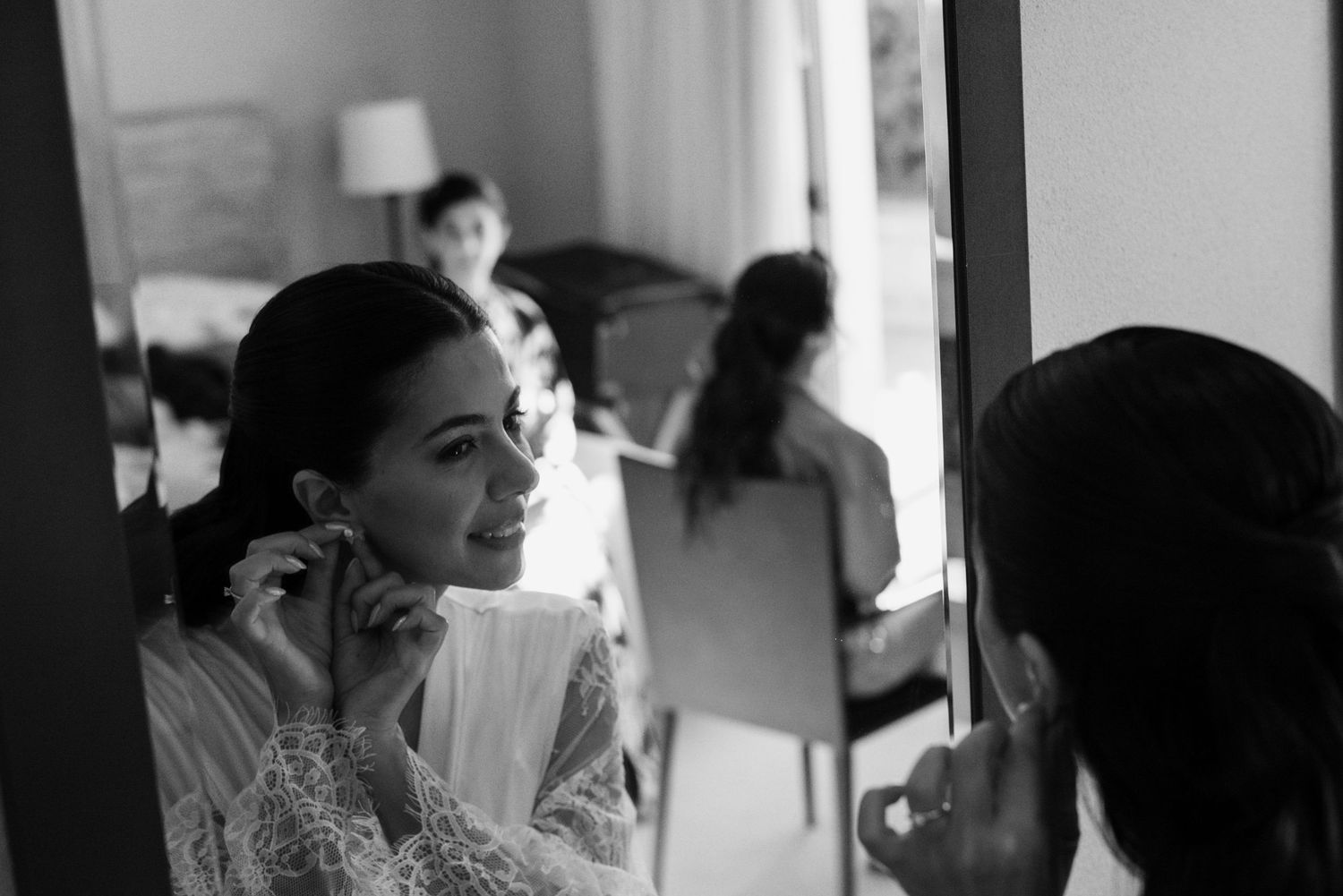 Bride adjusting earrings in mirror, bridesmaids in background, elegant black and white wedding preparation photo.