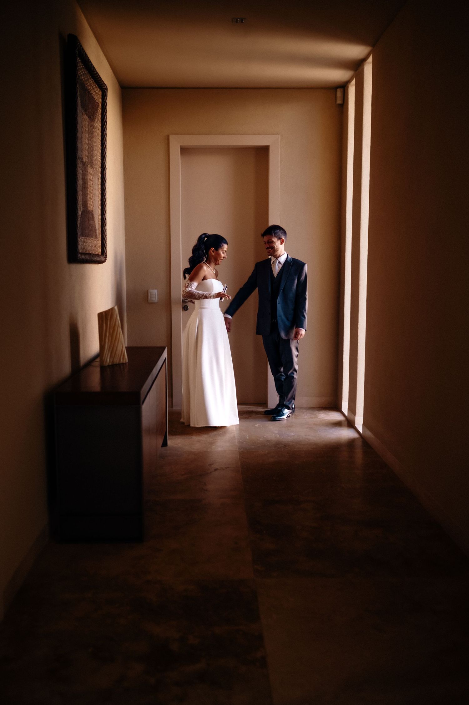 Bride and groom share a tender moment in a warmly lit hallway, bride in white gown, groom in navy suit.