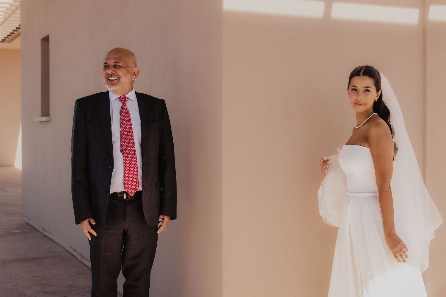 A bride in a white gown and veil stands beside an older man in a dark suit with a red tie at a sunny wedding venue.