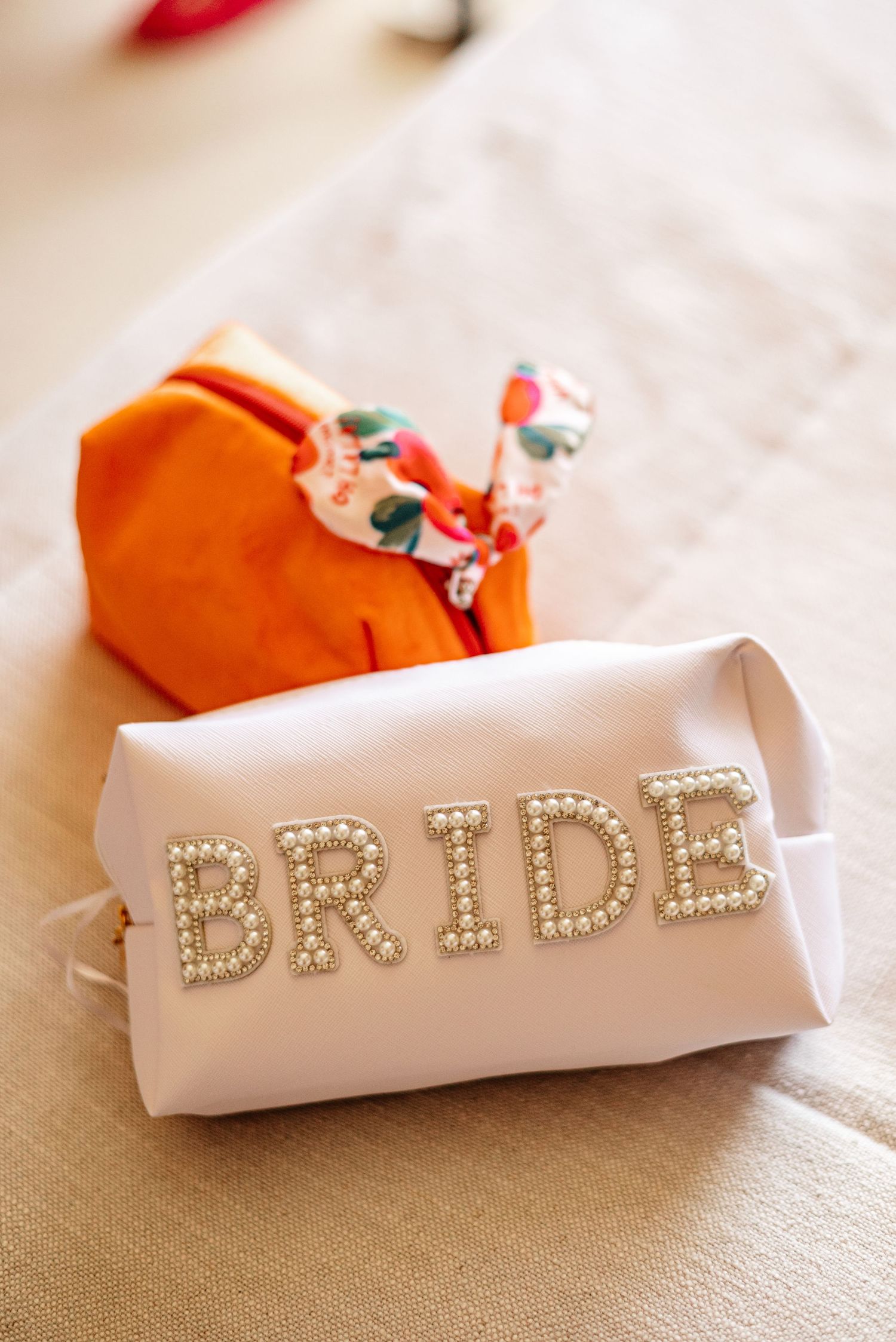 White pearl letter BRIDE cosmetic bag with orange gift wrap on neutral linen background for wedding day essentials.