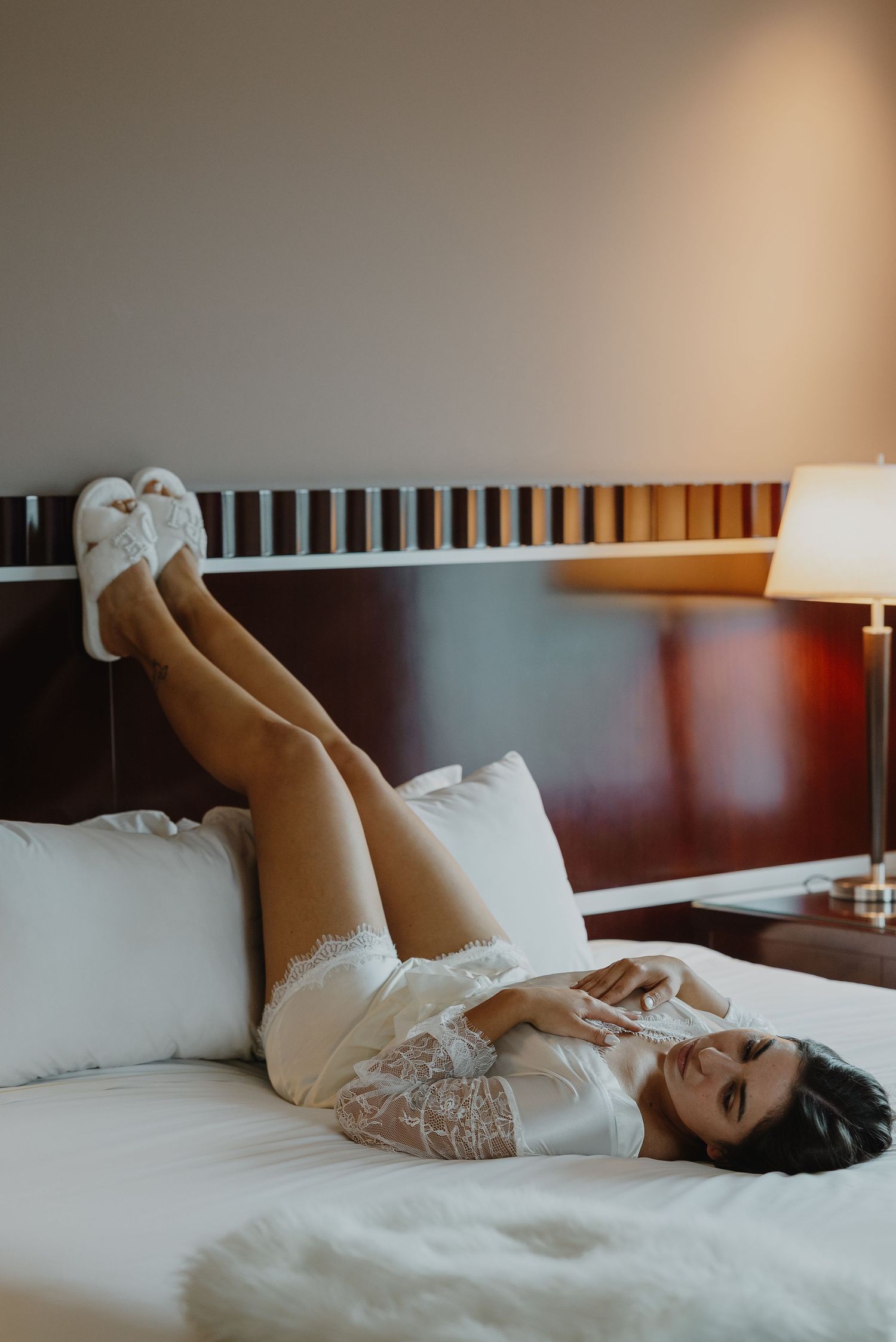 Woman in white lace robe and slippers lying on a hotel bed with legs raised, smiling in a cozy setting.