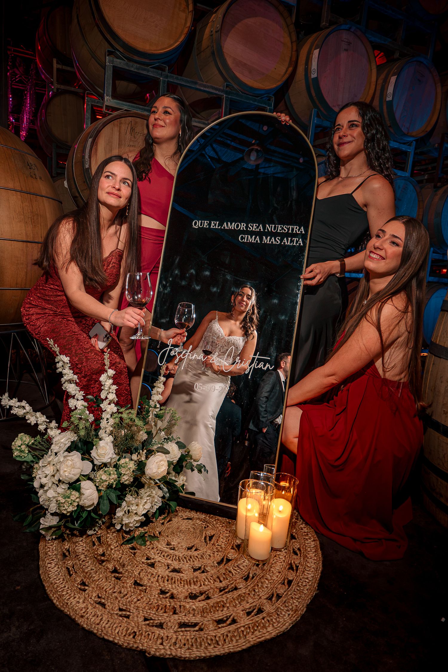 Bridesmaids gather around a decorative wedding mirror with candles and florals in a warmly lit winery barrel room.
