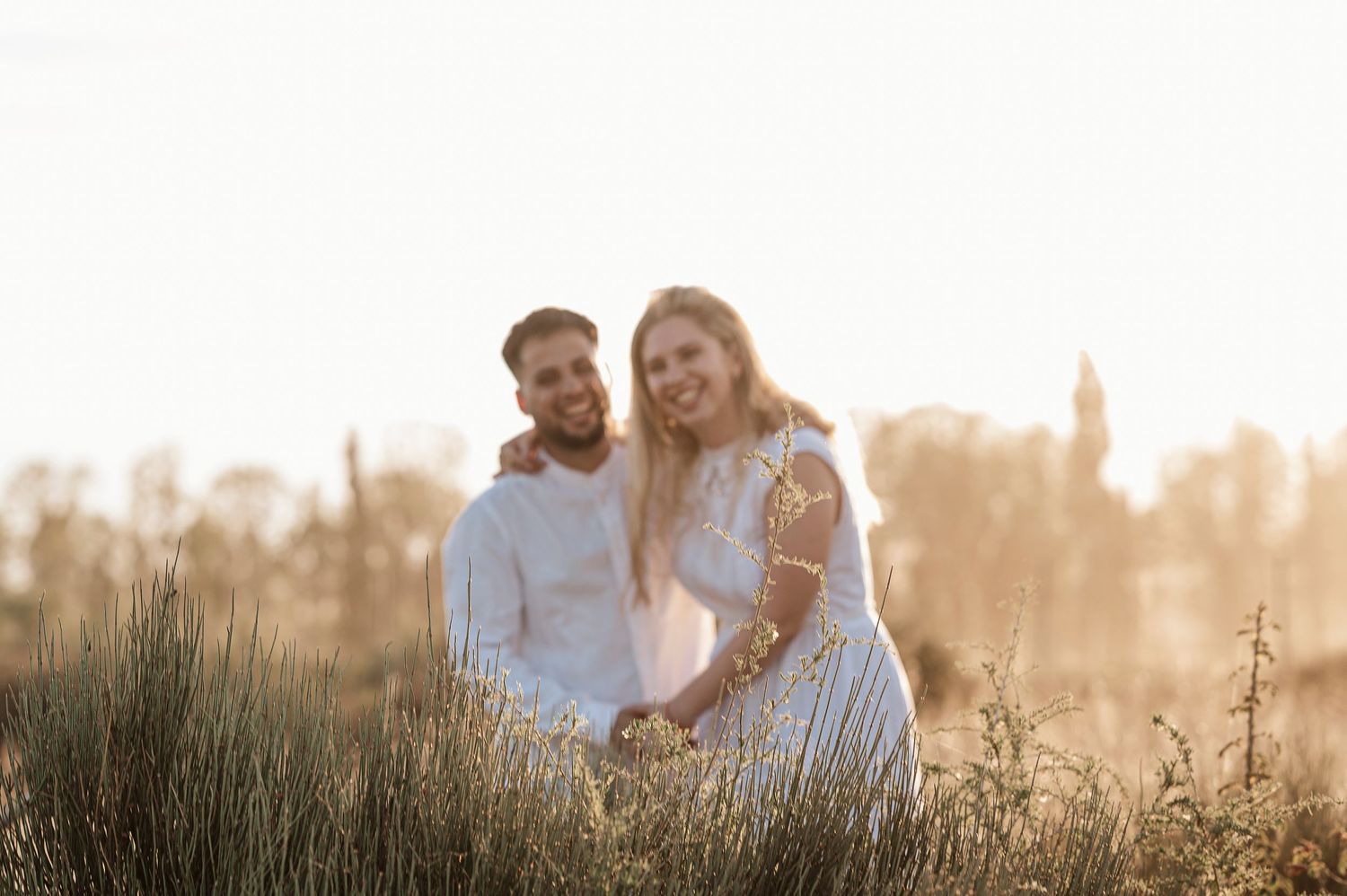 A couple sits together in a sunlit meadow with tall grass during golden hour, creating a romantic outdoor portrait.