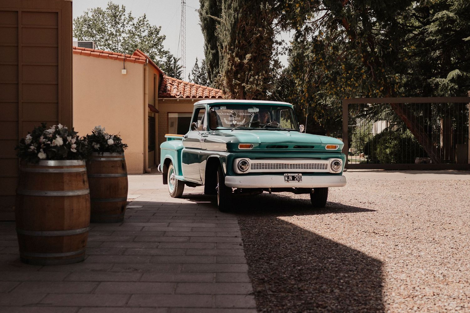 A vintage turquoise Chevrolet pickup truck parked on gravel next to wooden barrels outside a stucco building.