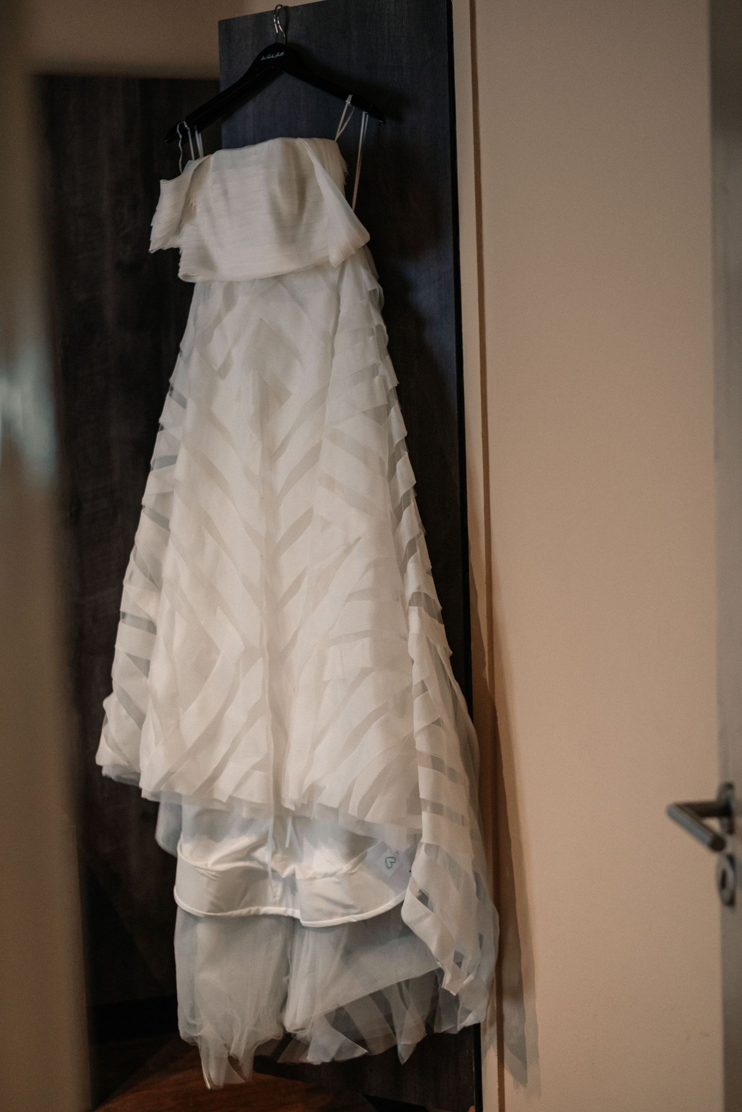 A white ruffled wedding dress with delicate layers hangs elegantly against a neutral wall.