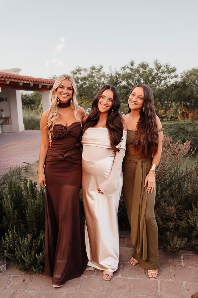 Three women pose together outdoors wearing brown, beige and olive colored dresses against a desert garden backdrop. wedding in mendoza