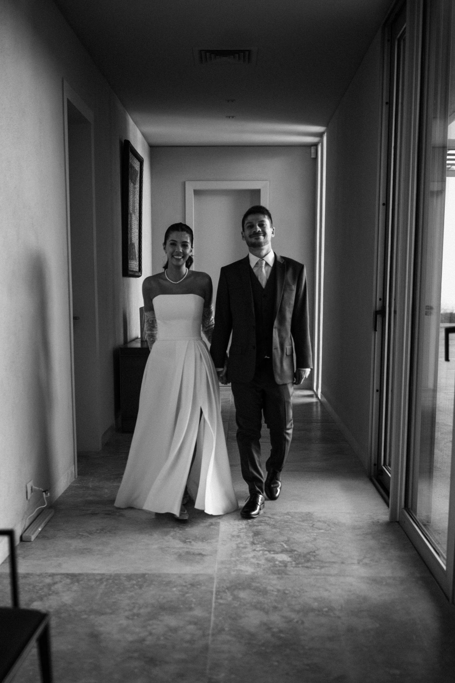 Black and white photo of a bride and groom walking hand in hand down a hallway on their wedding day.