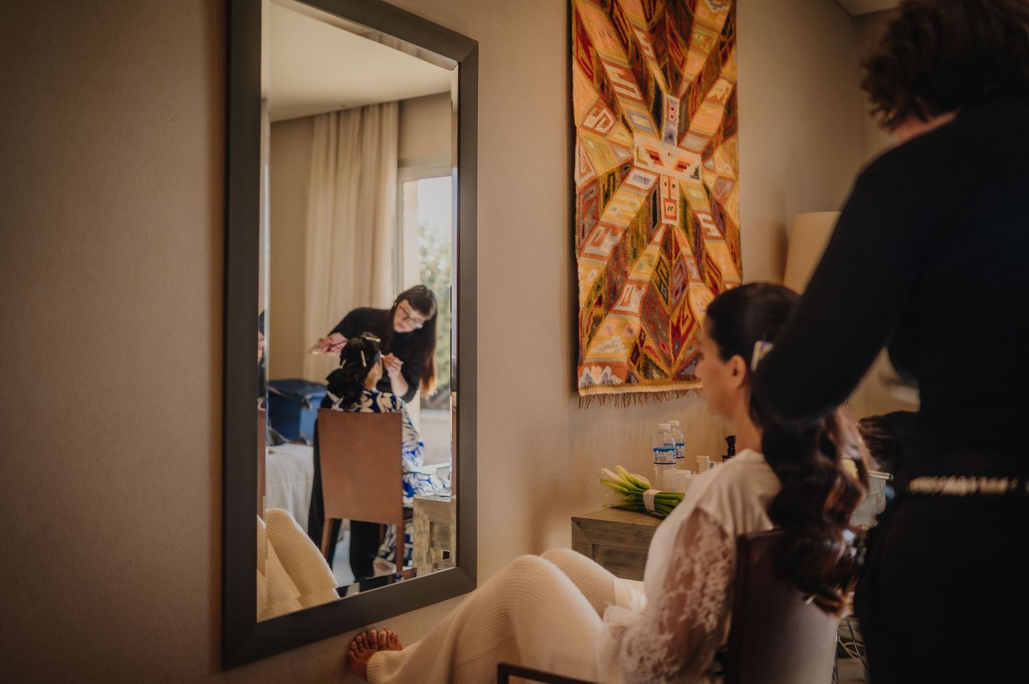 Bride getting hair styled in mirror reflection with wedding preparations in elegant hotel room.