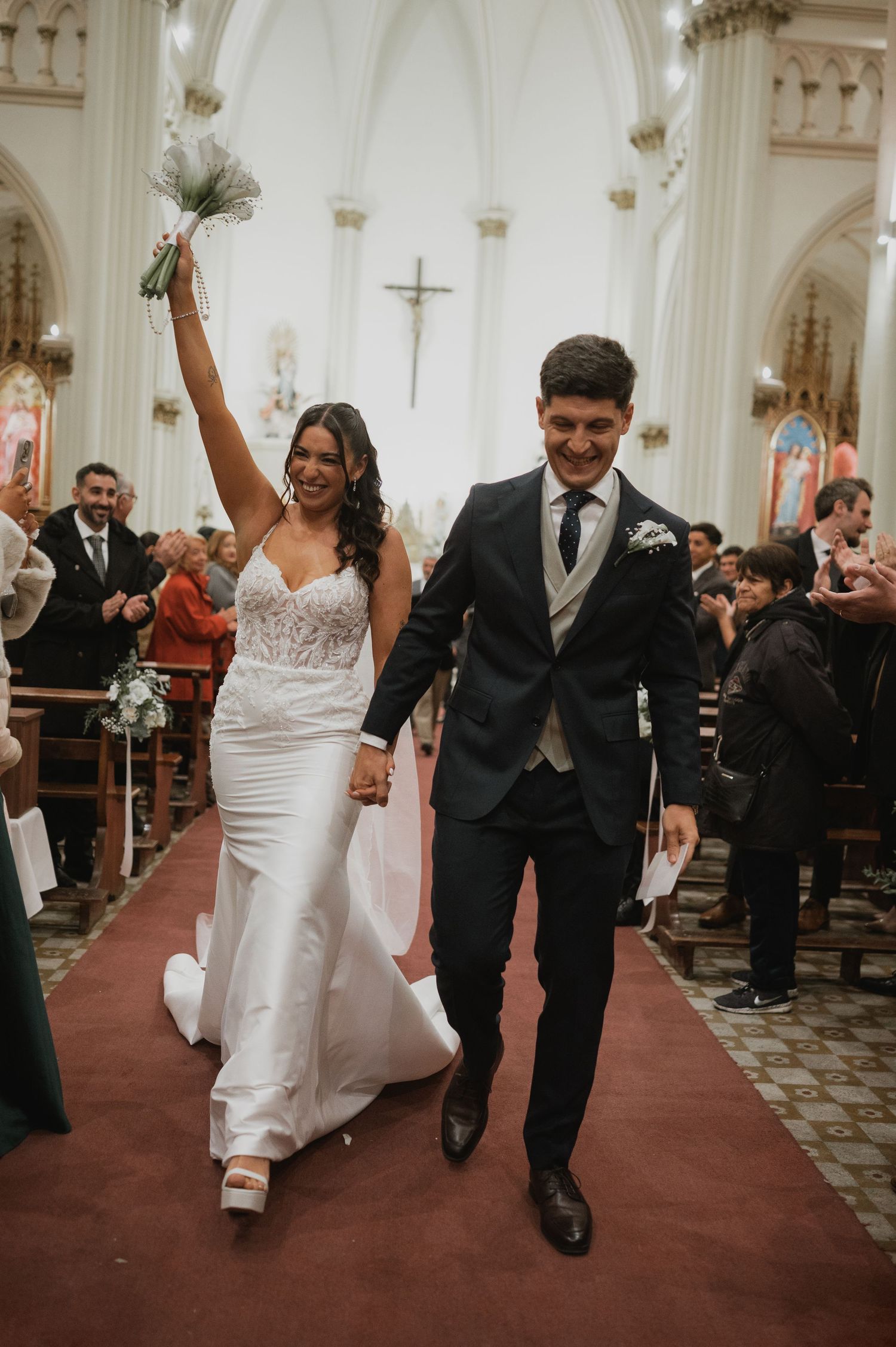 Joyful newlyweds walk down church aisle, bride in white gown raising bouquet, groom in dark suit, guests cheering.