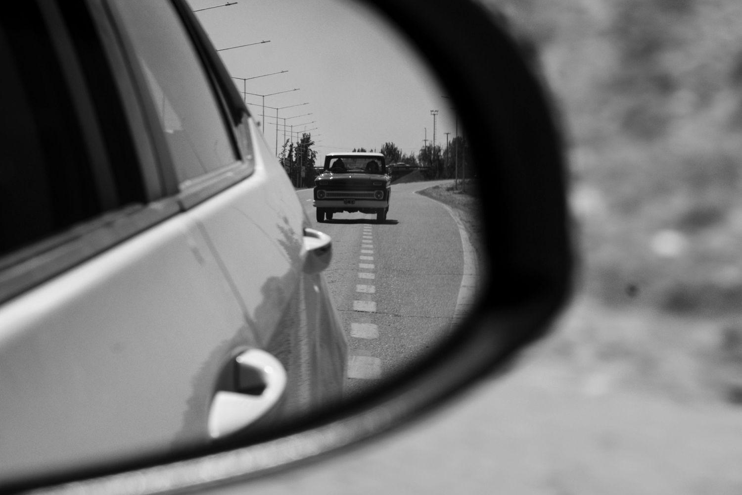 Car side mirrors show a series of trailing vehicles on a highway in black and white photography.