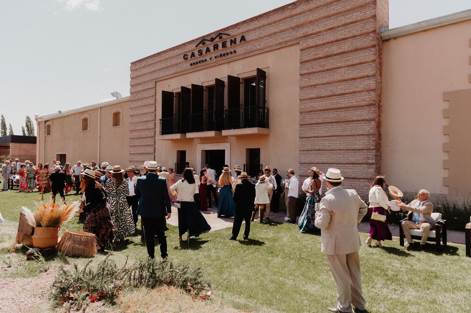 Guests gather outside a beige building for an outdoor event with multiple people mingling in groups.