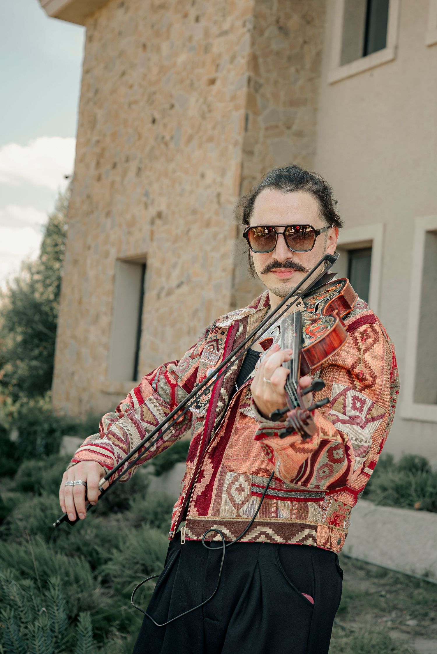 A musician plays violin while wearing a patterned sweater in a sequence of connected photos against a stone wall.
