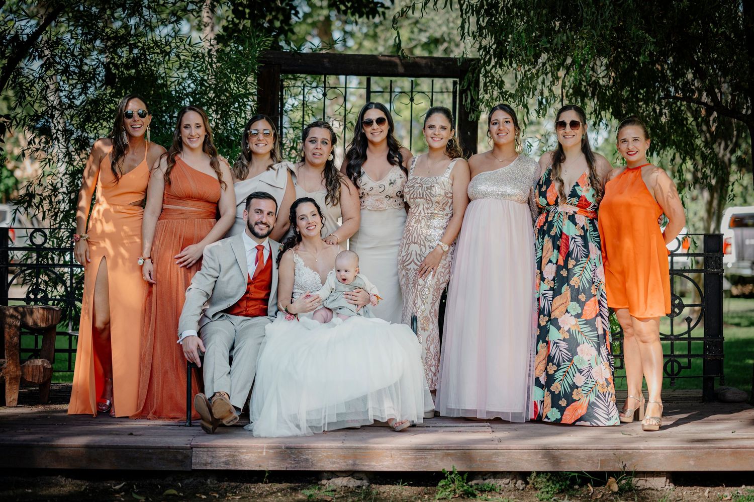 Wedding party members in warm peach, orange, and pink dresses pose together outdoors in a garden setting.