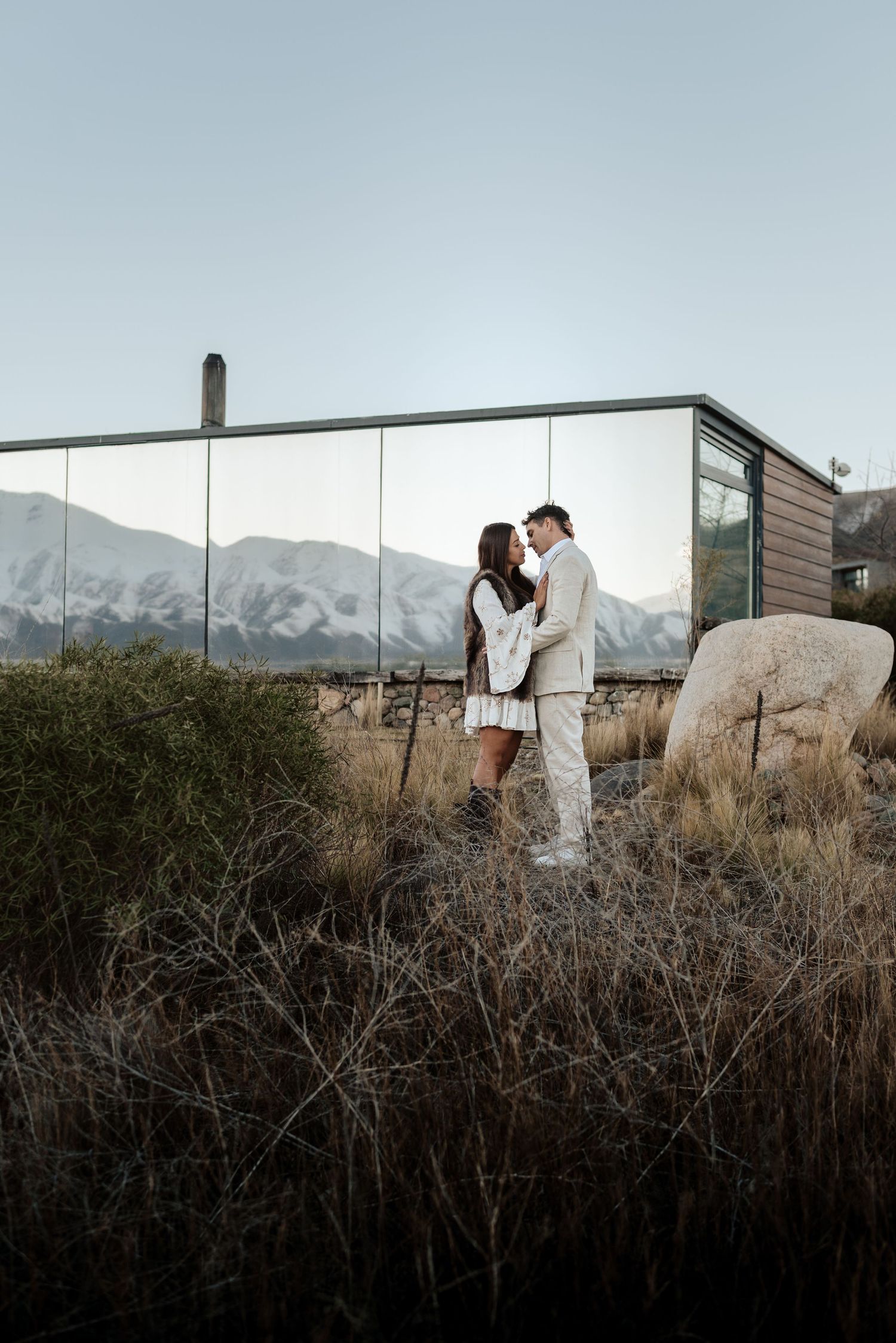 Una pareja se abraza frente a un edificio con espejos, vistas a la montaña y un paisaje desértico árido. pre boda mendoza