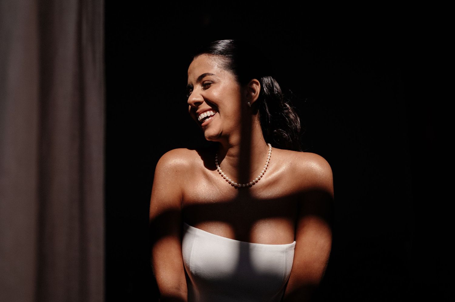 Smiling bride in strapless white gown and pearl necklace, bathed in dramatic light against dark background.