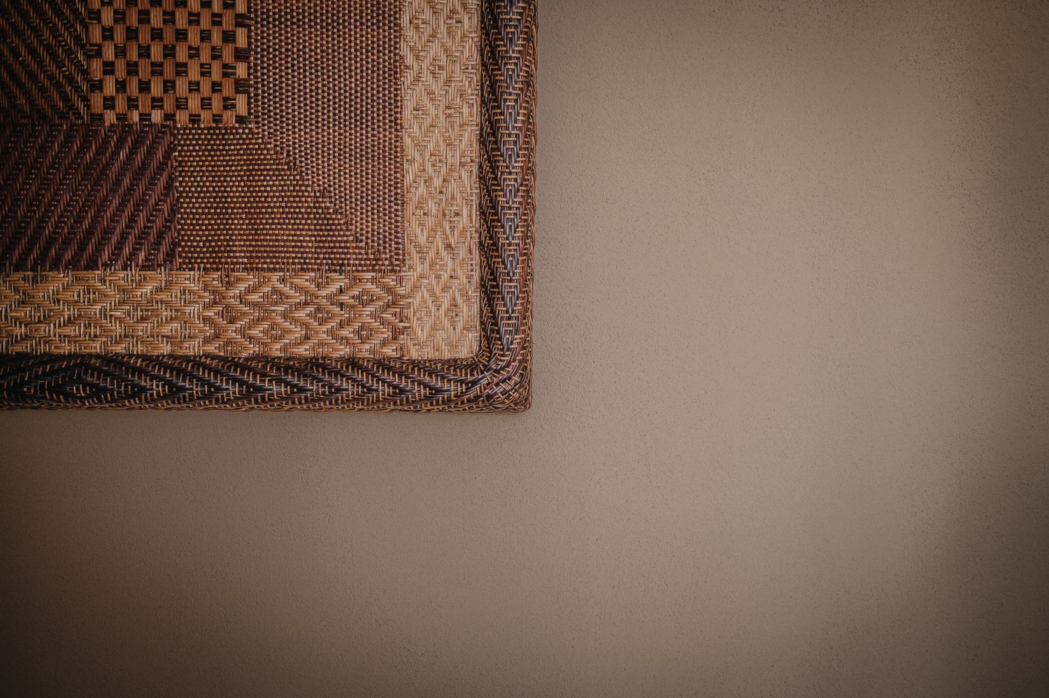 Corner of a decorative woven rug with geometric patterns in warm brown, tan, and burgundy tones against a neutral wall.