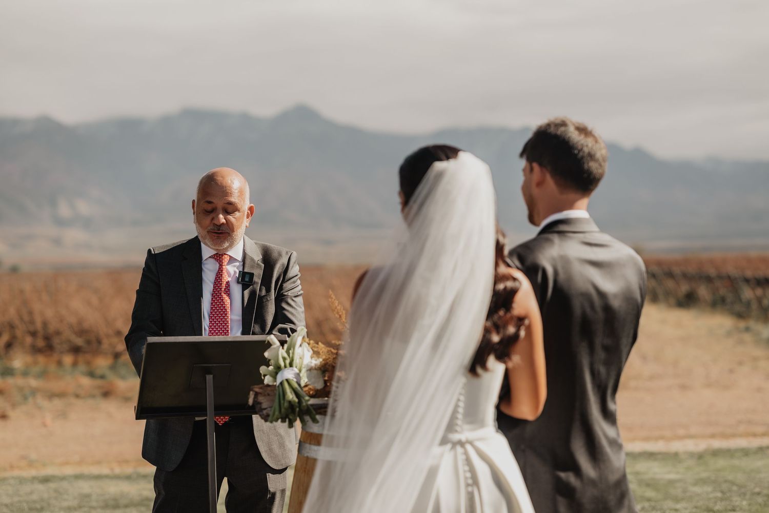 Outdoor wedding ceremony with officiant at podium, bride in white gown and veil, groom in gray suit, mountain backdrop.