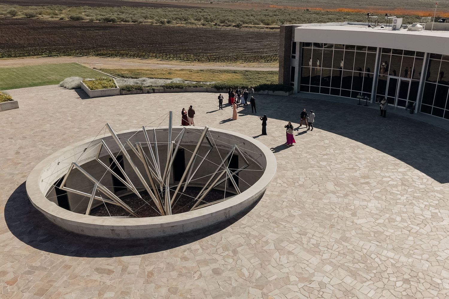 Aerial view of a modern museum entrance with a circular sunken sculpture installation on a stone plaza in an arid landscape. Mendoza